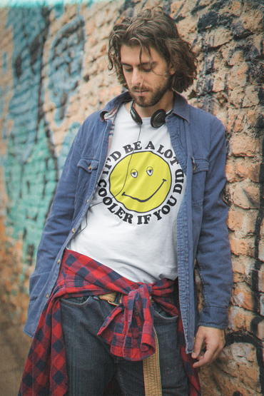 Man in casual clothes with a smiley face t-shirt leans against a graffiti wall.