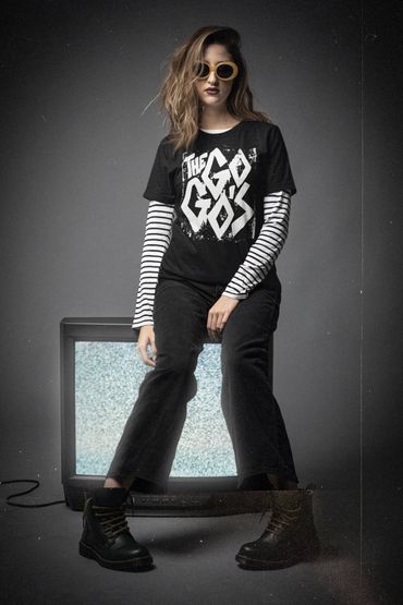 Woman with yellow sunglasses sitting on a TV showing static, wearing a black band tee and striped long sleeves.