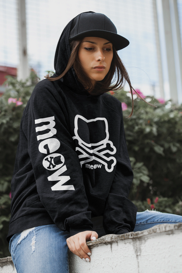 Young woman in a black hoodie with a cat and crossbones design, wearing a black cap.