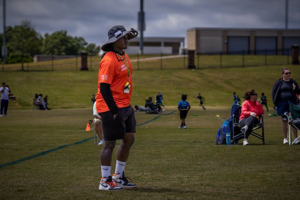Coach in an orange shirt and hat watches a youth sports game on a grassy field.