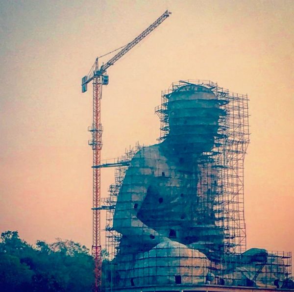 Large statue under construction with scaffolding and crane at sunset.
