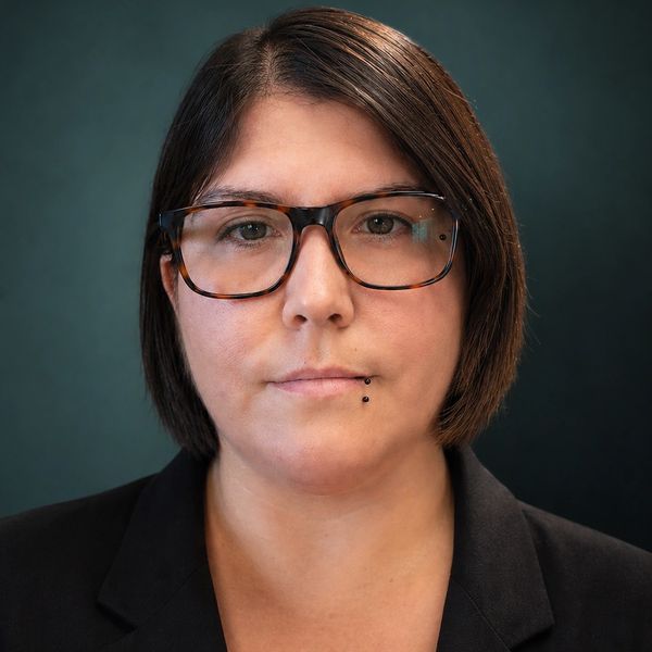 Serious woman with glasses and lip piercing against a dark background.