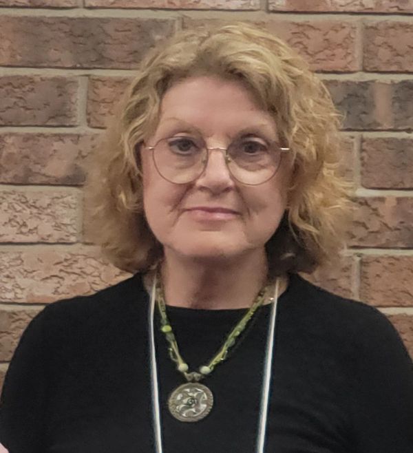 Smiling woman with glasses and curly hair in front of a brick wall.