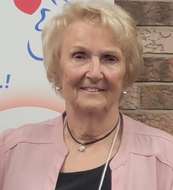 Smiling elderly woman with short blonde hair wearing a pink jacket and necklace.