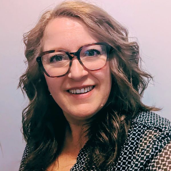 Smiling woman with curly hair and glasses wearing a patterned top.