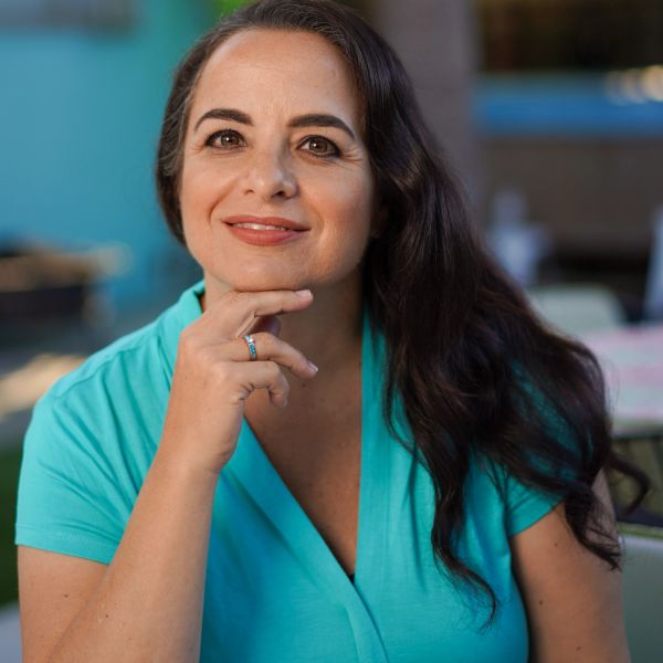 Smiling woman with long dark hair and turquoise top, resting her chin on her hand.