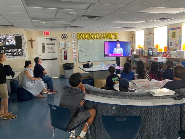 Students and adults watch a presentation in a classroom with 'ROBOCANES' balloons.