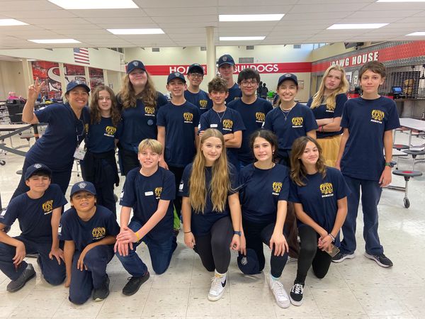 Group of students and a teacher in matching navy blue shirts posing in a school cafeteria.