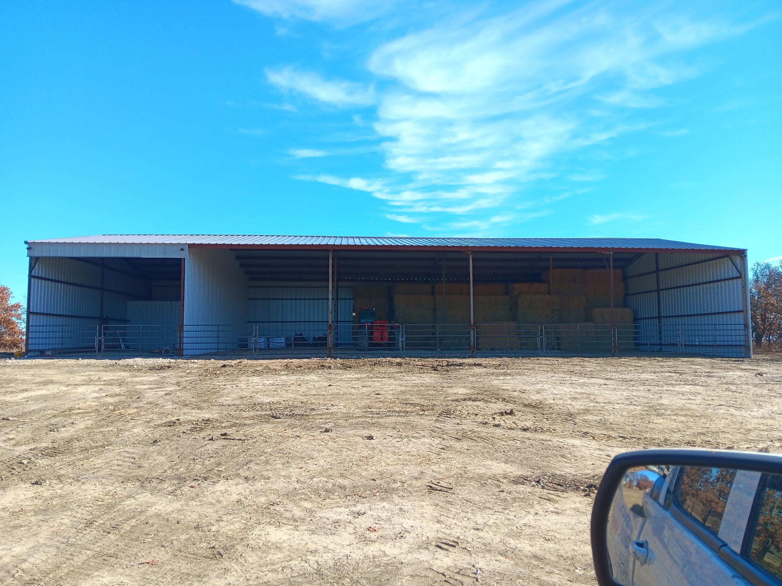 Open metal barn storing hay bales and farming equipment under a clear blue sky.
