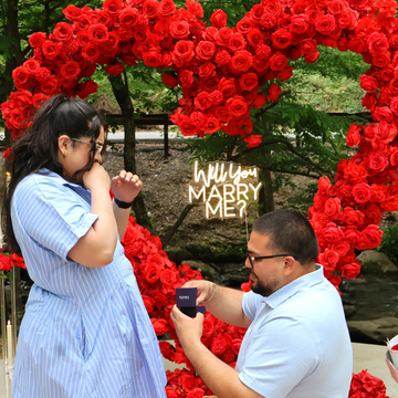 Man proposing to woman in front of a heart-shaped rose backdrop.
