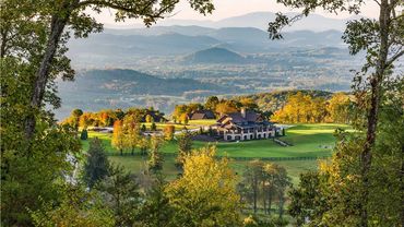 A large house surrounded by green fields and colorful autumn trees in a mountainous landscape.