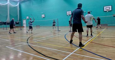 Group of men playing badminton indoors on wooden court with basketball hoops.