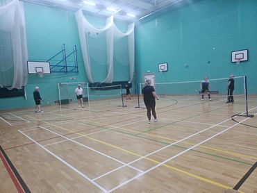 Adults playing badminton in an indoor sports hall with teal walls.