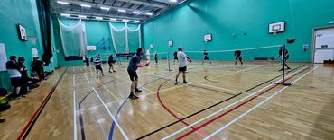 People playing badminton in a spacious indoor gymnasium with green walls.