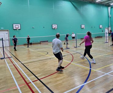 Group of adults playing badminton indoors in a gymnasium with basketball hoops.