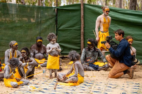 Indigenous people in yellow paint and skirts participating in a cultural event while a cameraman records.