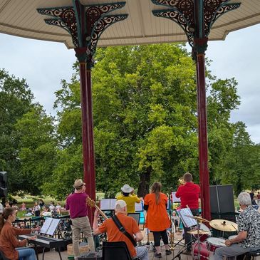 Band performs outdoors under a decorative pavilion in a park.