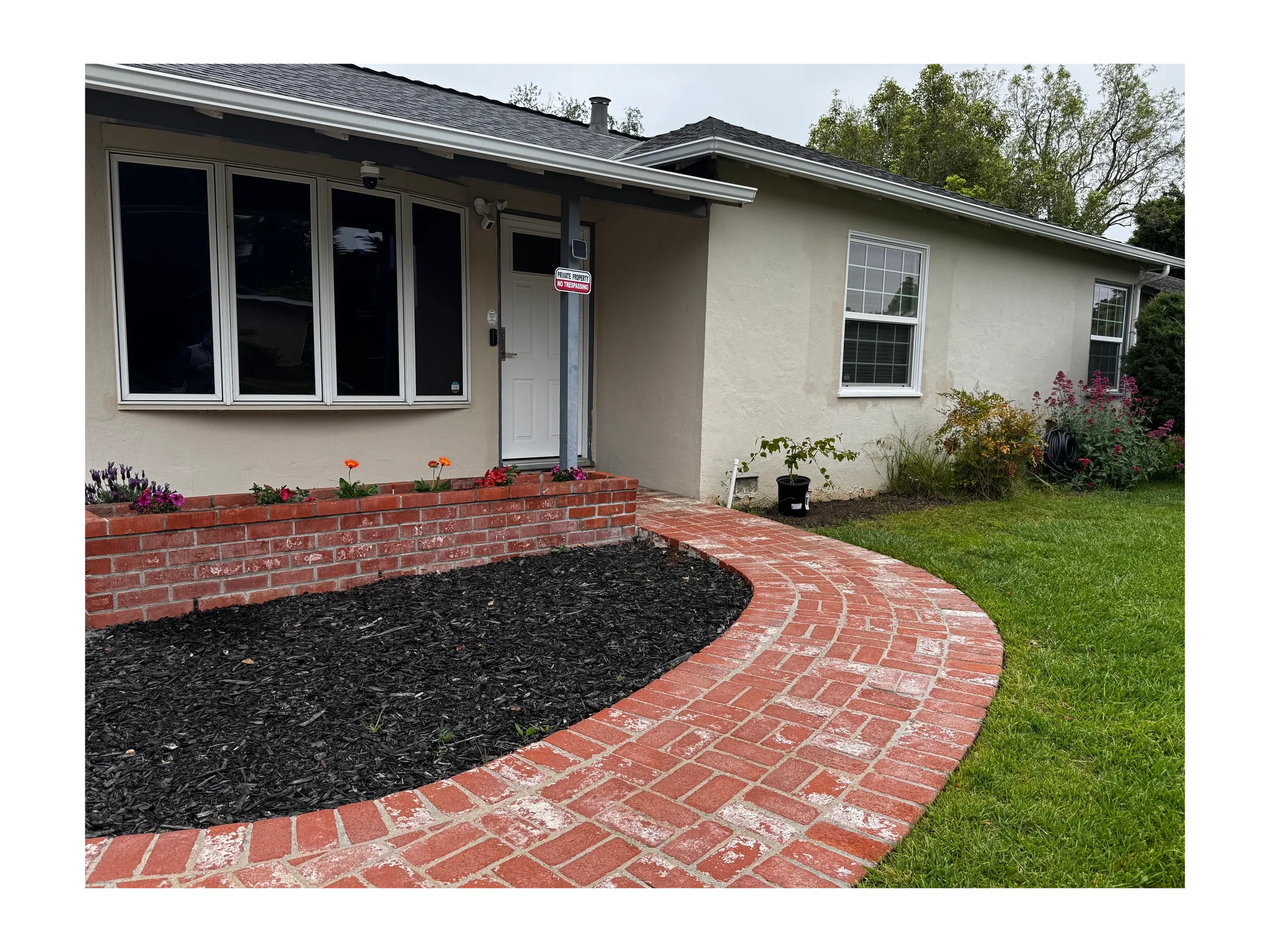 Curved red brick pathway leading to a house entrance with flower beds and greenery.