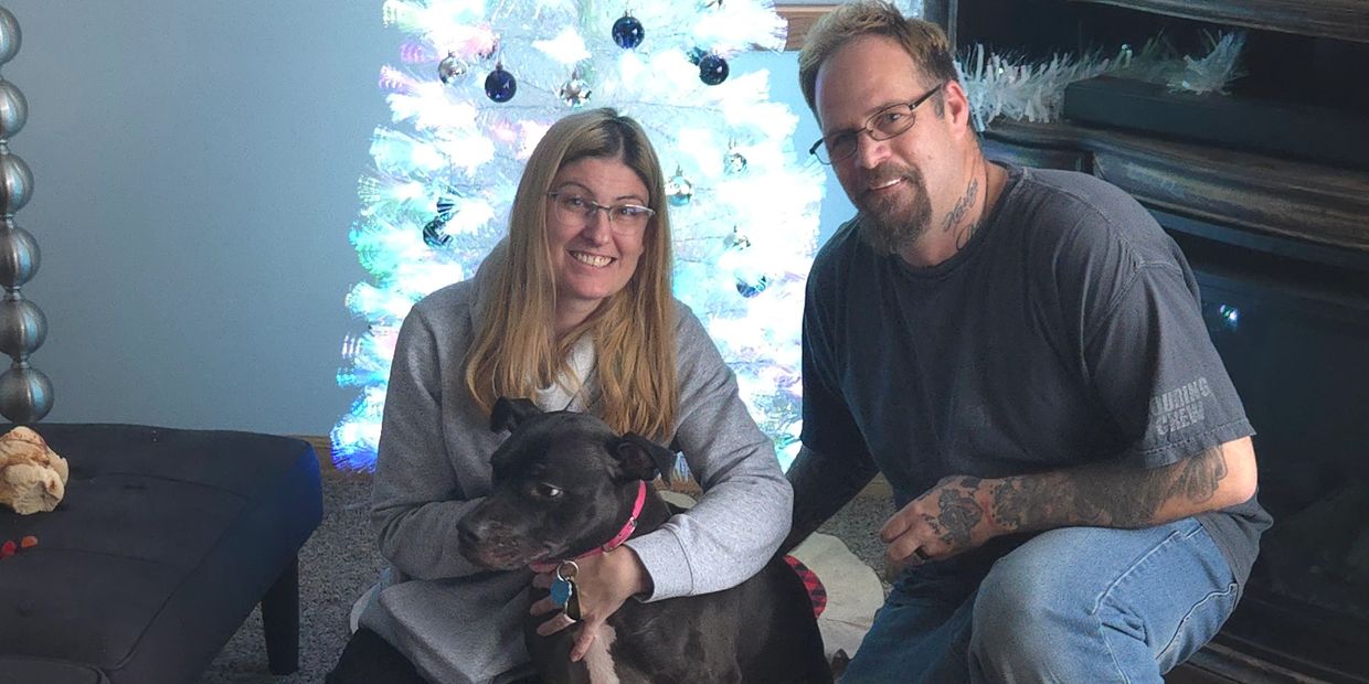 Smiling couple with their black dog in front of a white Christmas tree.