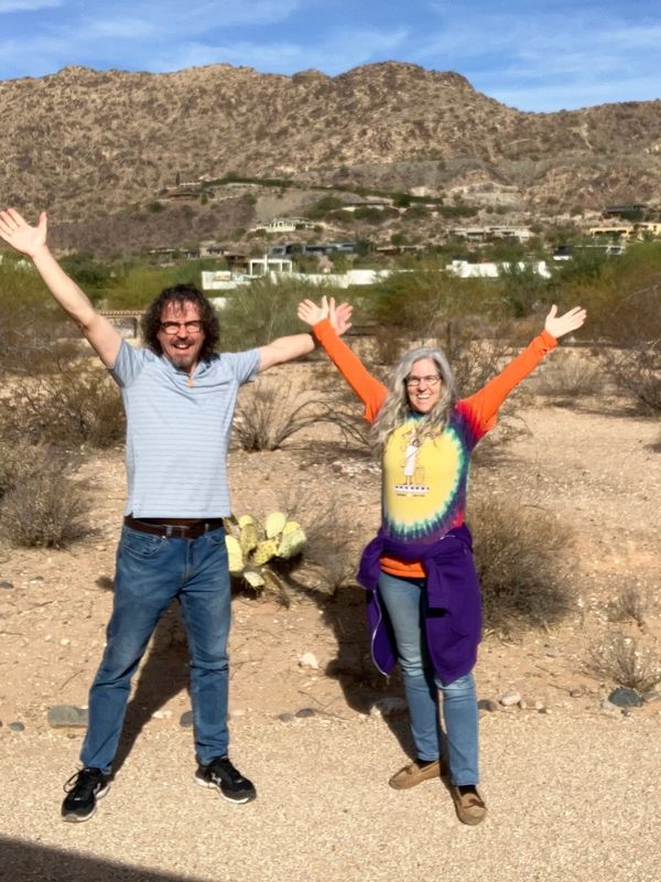 Two people joyfully raise their arms in a desert landscape with mountains in the background.