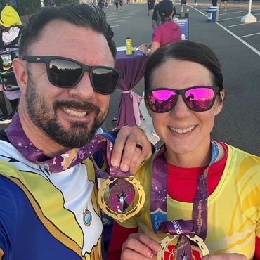 Smiling couple proudly displaying their race medals at an outdoor event.