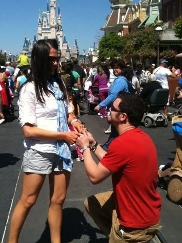 Man proposing to woman at a busy amusement park street with a castle in the background.