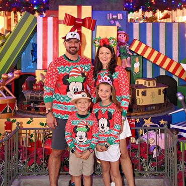 Family in festive Mickey and Minnie Christmas sweaters posing under a decorated tree with gifts.