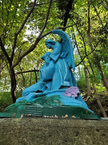 Blue angel statue comforting a kneeling figure in a forest.