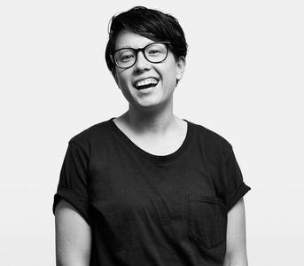 A black and white photo of Emily Hashimoto, They're smiling, wearing glasses and a black T-shirt.