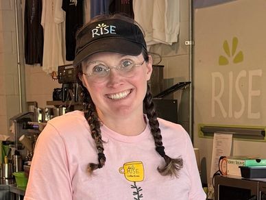 Smiling barista wearing a pink t-shirt and black Rise cap behind a coffee counter.