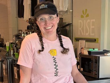 Smiling barista wearing a pink t-shirt and black Rise cap behind a coffee counter.