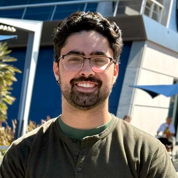 Man with glasses smiling outdoors in casual attire.