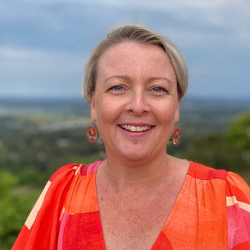 Smiling woman in a vibrant orange dress outdoors with a scenic background.