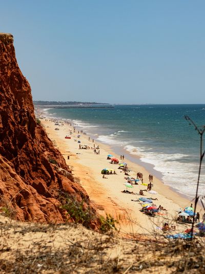Tan sand with many sunbathing people on it, with large red cliffs behind and the ocean in front