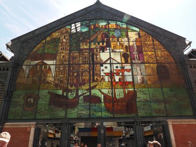 Stained glass mural depicting boats and historic buildings over a market entrance.