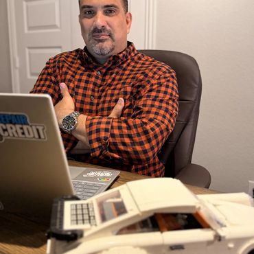 Man in checkered shirt sitting at a desk with a laptop and a white model car.