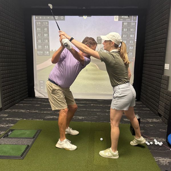 A woman instructs a man on his golf swing in an indoor simulator.