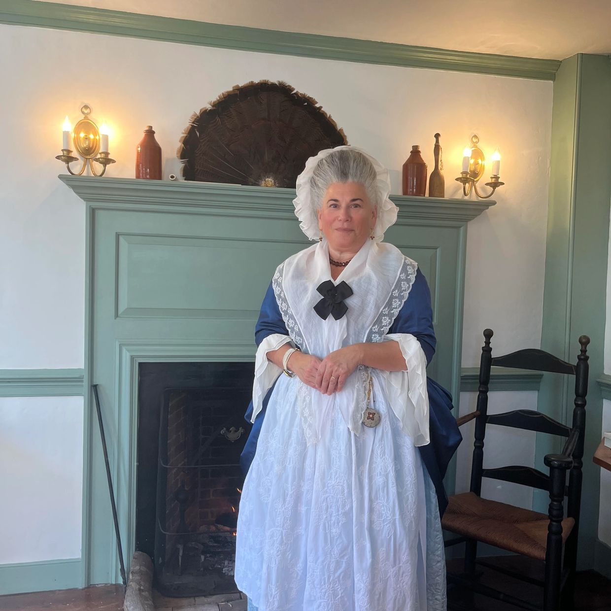 Sandy Spector as Martha Washington in a sapphire blue silk satin gown in front of a fireplace.
