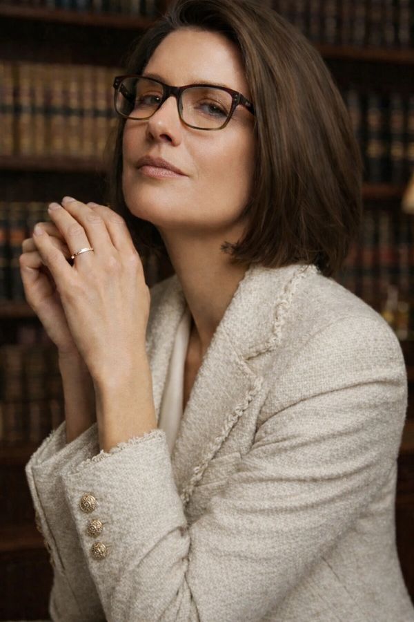 Woman in glasses and beige blazer posing thoughtfully.