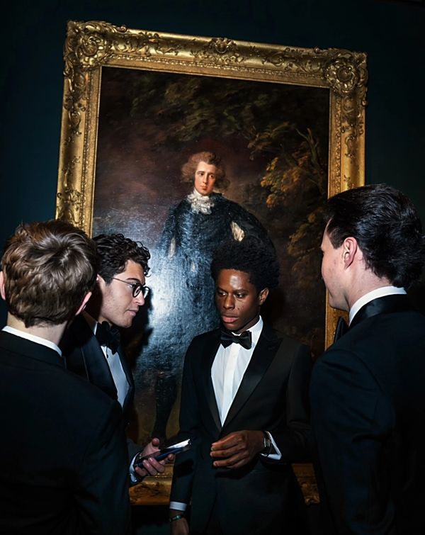 Four men in tuxedos talk in front of a classical portrait.