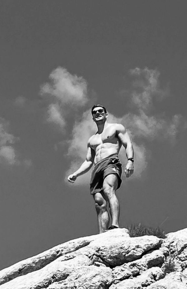 Shirtless man standing on a rocky peak against a cloudy sky.