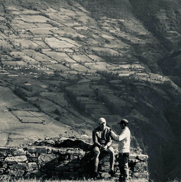 Two men interacting on a stone structure against a mountainous backdrop.