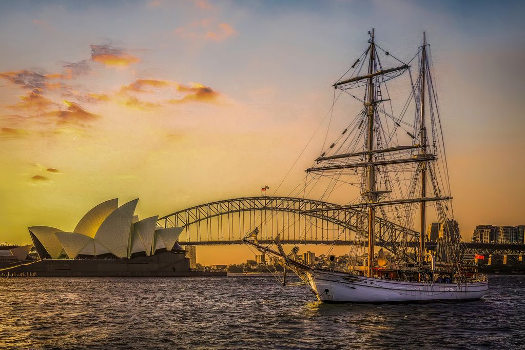 Sailing ship near Sydney Opera House and Harbour Bridge at sunset.