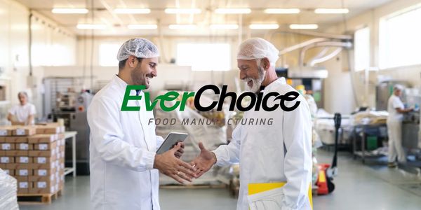 Two food industry professionals in white coats and hairnets shaking hands in a manufacturing plant.