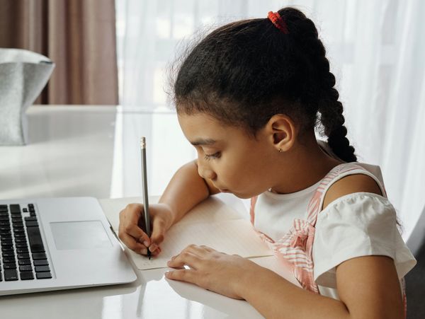 Young girl focused on writing homework beside a laptop.