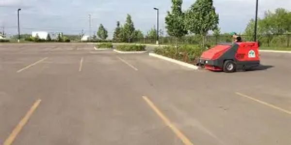 A person operates a red street sweeper in an empty parking lot on a clear day.