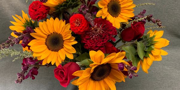 A vibrant bouquet with sunflowers, red roses, and purple flowers on a gray fabric background.