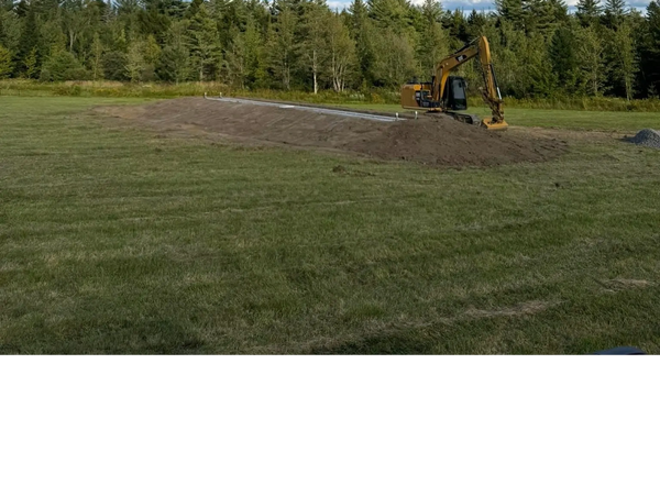 Excavator working on a mound of earth in a grassy field with trees and blue sky. Another septic system going in!