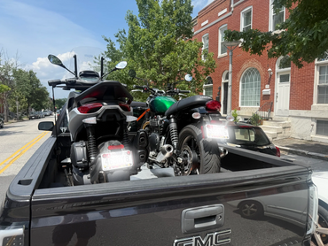 Two motorcycles loaded in the back of a black GMC pickup truck on a sunny street.
