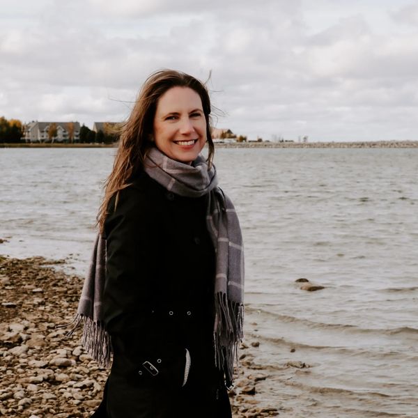 Woman smiling and wearing a jacket and scarf at a shoreline with houses in the background